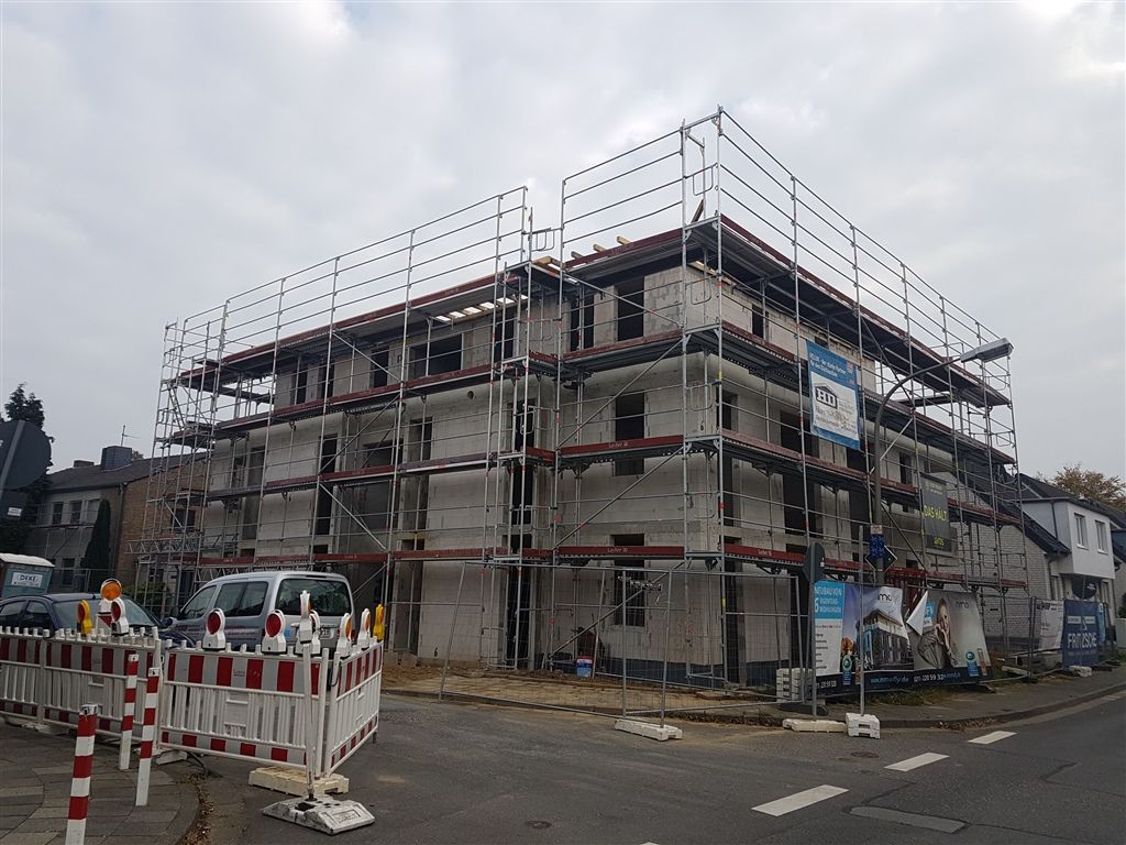 Rohbau eines Mehrfamilienhauses hinter einem Gerüst an einer Straßenecke, Nachbarhäuser daneben.